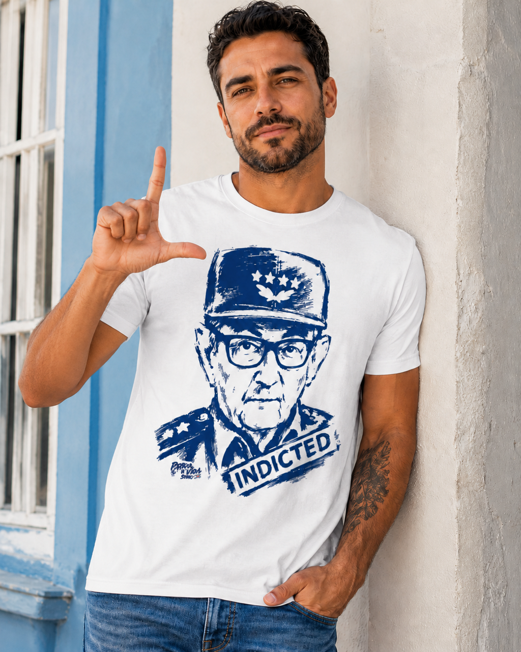 Man wearing a white t-shirt with a graphic of a person in a military uniform and the word 'INDICTED' pointing at a blue wall.